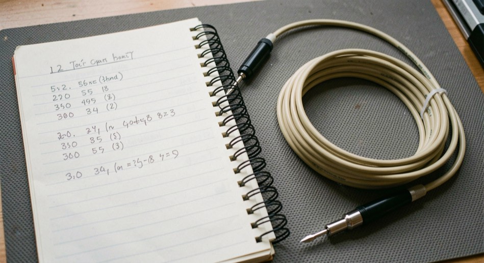 A spiral notebook open beside a probe tip and a coil of coaxial cable on a grey bench mat, handwritten measurements filling the left page